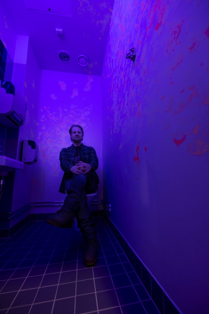 Mies istuu UV-valaistun WC:n pöntöllä.
