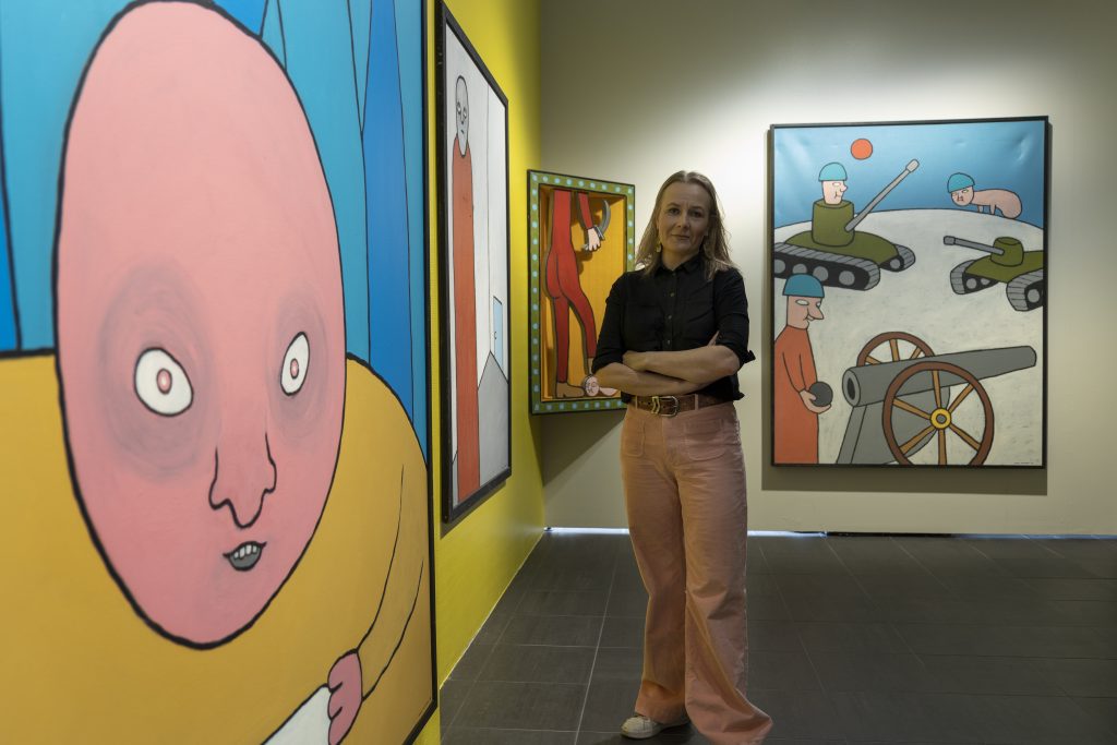Nainen seisoo kädet ristissä taidemuseon näyttelytilassa, jonka seinillä on Mikko Valtosen värikkäitä teoksia.