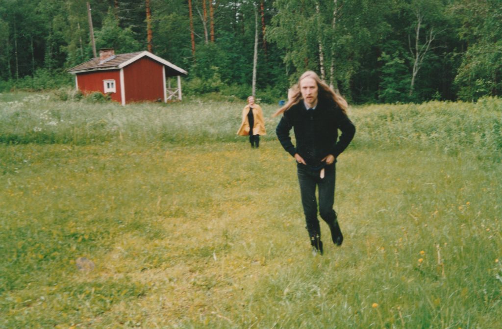 Kesäinen maisema, jossa on kaksi henkilöä. Vihreällä ruoholla kävelee mustiin pukeutunut pitkähiuksinen mies. Taaempana näkyy toinen ihmishahmo. Taustalla on punainen mökki.