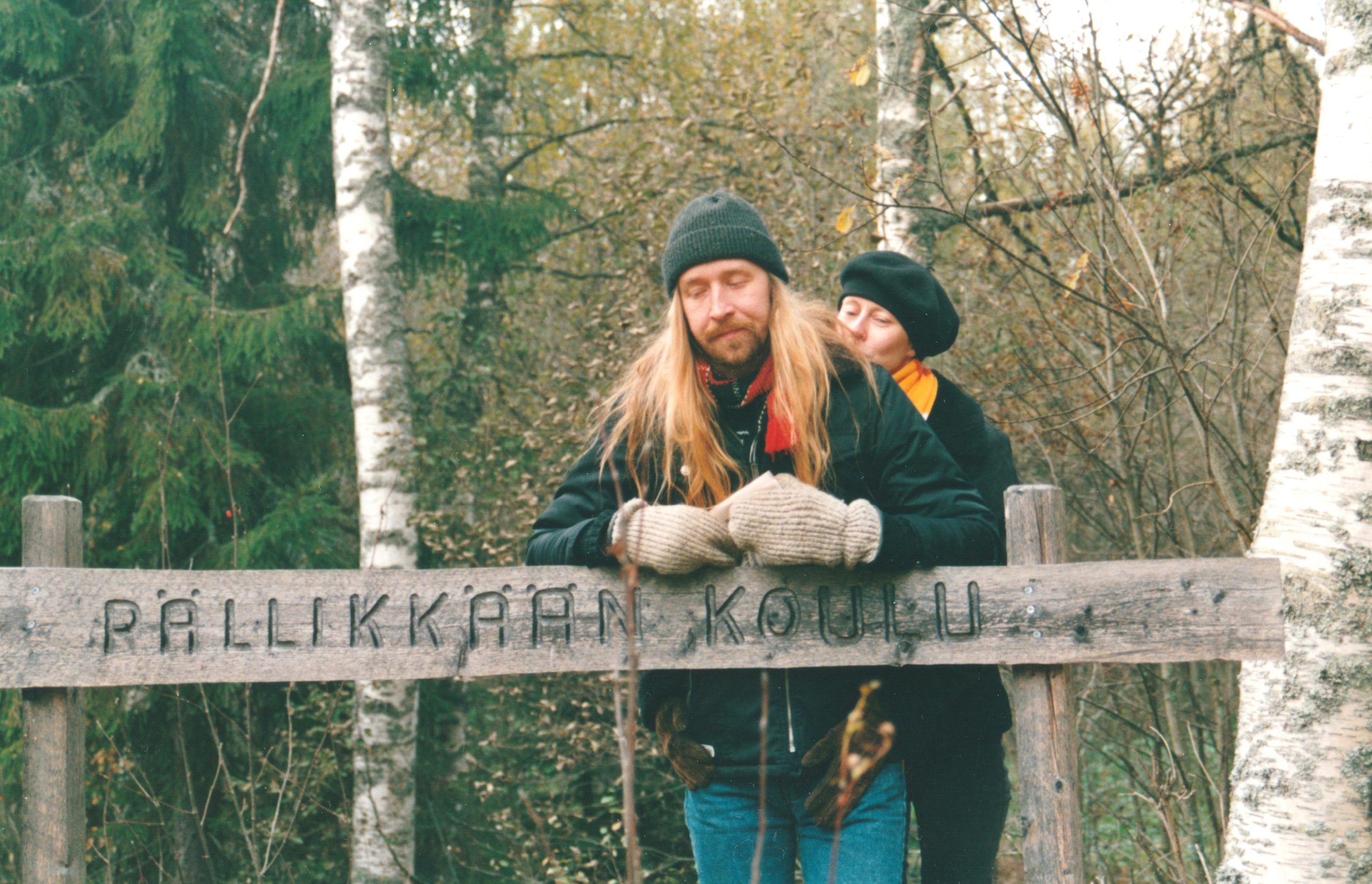 Kaksi henkilöä nojaamassa puiseen kylttiin. Kyltissä lukee "Pällikkään koulu". Etummaisena pitkähiuksinen mies. Hänen takanaan mieheen nojaa nainen, kädet hänen ympärillään.