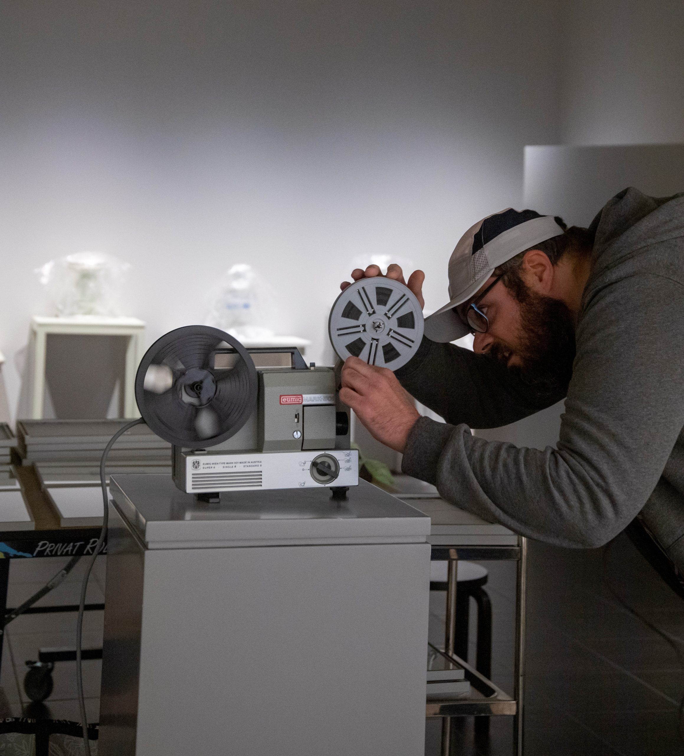 Lippalakkipäinen mies asettaa elokuvafilmikelaa jalustalla seisovaan kaitafilmiprojektoriin.