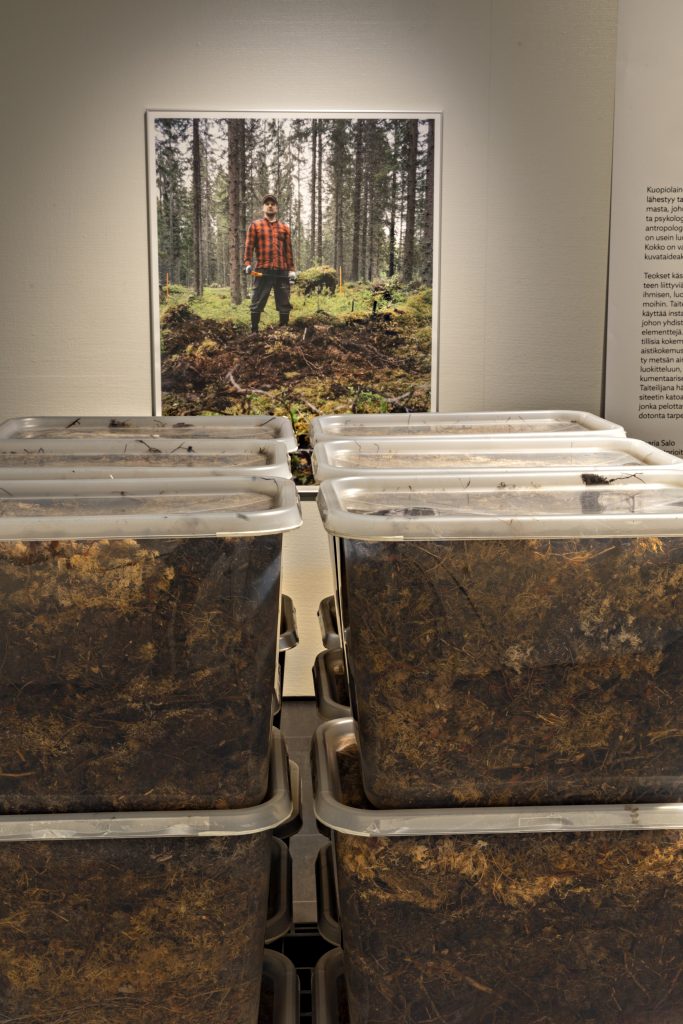 Etualalla on kasassa läpinäkyviä muovisia laatikoita, joiden sisällä on metsän pohjakerrosta. Muovilaatikoiden takana seinällä on kuva ruutupaitaisesta miehestä, joka seisoo metsässä lapio kädessä. Miehen ympäriltä maata on kaivettu, ja lapiossa on ylös kaivettua maata ja sammalta.