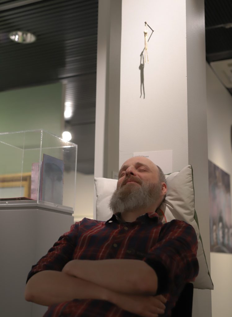 Harmaapartainen ruutupaitainen mies nojailee istualtaan tyynyyn teoksen alla silmät kiinni.